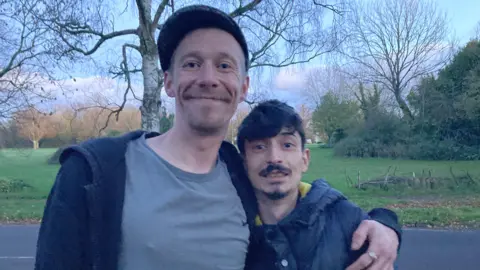 Two men posed for a picture in front of a park. The taller man on the left is Neville, who is wearing a grey t-shirt, black hoodie and a hat. He has his arm around Tyler, who is shorter and has dark-brown hair and is wearing a black coat.