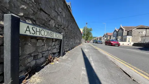 BBC A residential road with a sign saying Ashcombe Road on it, and cars in the background