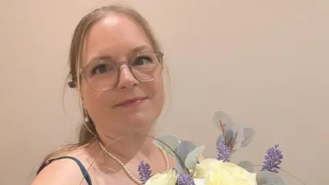 Ruth Hayman A woman with blonde hair and glasses, wearing pearl earrings and a pearl necklace, is holding a bouquet of flowers.
