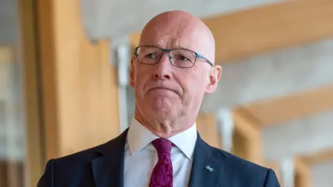 PA Media John Swinney walking down a Holyrood corridor - he is a bald-headed man with glasses, a dark suit, purple tie and a serious expression on his face.