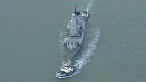 HMS Dragon is pulled out of Portsmouth harbour by two tug boats.