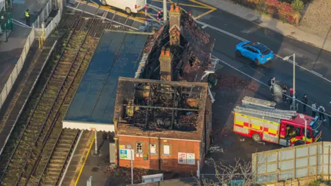 Oliv3r Drone Photography An aerial shot shows damage to the roof of the building, including charred wooden beams. Almost all of the rood has been destroyed. A fire engine is next to the building and police tape is in place. The railway line is visible on the left hand side of the photo. 