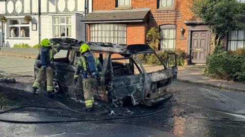 Two firefighters are dealing with the burnt carcass of a campervan in the middle of a road. There are buildings in the distance. There are people sat in the window of one building which is painted white. There is a bay window on another brick-built building adjoining it.