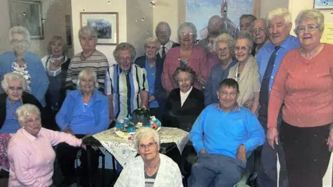 More than 20 elderly men and women are gathered round for a group photo in the BRICC house. 