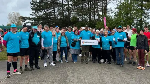 Ryan O'Shea A large group of participants wearing matching turquoise shirts stand together on a paved path at an outdoor park, posing for a group photo during a charity running event, with trees and other runners in the background.