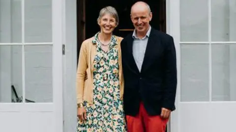 Christian Michael The picture shows two people standing side by side in the open doorway of a large, traditional house. The doorway is painted white and made up of multiple glass panes. Both people are standing on a stone step, facing the camera. One person is wearing a long, patterned dress with a light‑coloured cardigan. The other person is wearing a dark jacket, a light shirt and bright red trousers.