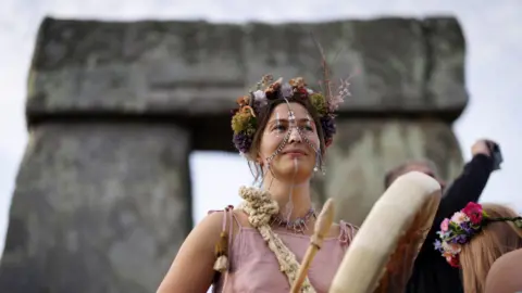 A woman in a flower crown and wearing jewellery chains over her face plays a handheld drum. An archway of the Stonehenge stones is behind her.