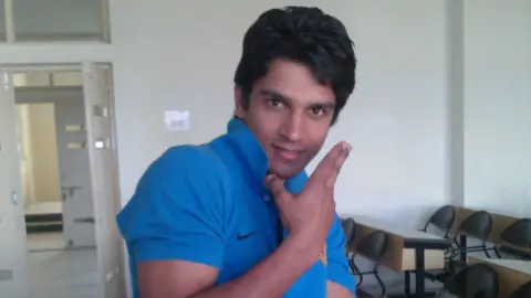 A young man wearing a blue T-shirt strikes a pose and smiles while looking at the camera in a college classroom