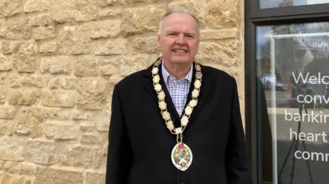 A man wearing a black suit, a white and purple checked shirt and a mayoral chain standing in front of a building 