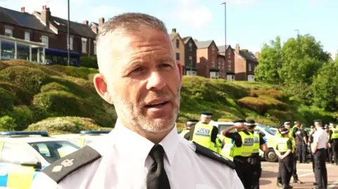 Chief Superintendent Jamie Henderson - who has grey sort hair and a grey beard wearing a police unform. There are police officers and police vehicles behind him.