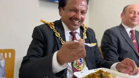 Jon Wright/BBC Elango Elavalakan wearing his mayoral chains and a suit, eats food at a table