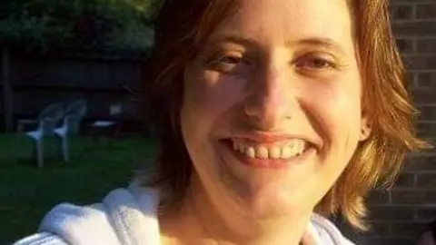 Juliet A woman dressed in a white hoodie smiles at the camera. He face is lit up by the sunshine and she has a brown shoulder-length hair.