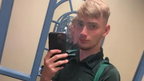 Facebook Jamie Kadolski stands in front of the mirror and takes a selfie. He has a black phone next to his face and looks directly into the mirror. He's wearing a green shirt. 