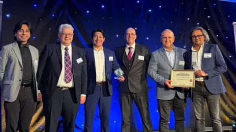 University of Essex A group of men with smart shirts and blazers on, smiling and holding up a framed certificate