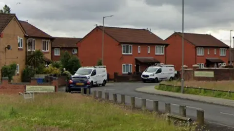 Three semi-detached brick houses on a street which has a grass area to the side. A number of stone bollards line the pavement and two white vans and a small black car are parked in the street.