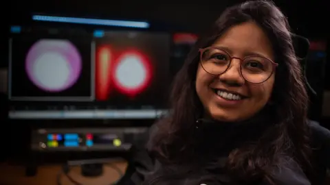 Tony Jolliffe/BBC News A woman, Veronica Viera, is sitting in front of two large computer monitors in a dark room. The screens show two images of a white and red circular shape - this is plasma glowing in the space furnace. Veronica has long brown hair, she is wearing glasses and a black jacket.