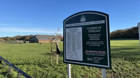 George Carden / BBC The sign for the recreation ground car park in the foreground with terms and conditions on it. In the background is the park and sports centre which cricket and football teams use