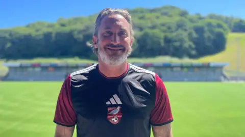 BBC/Peter Spencer Scarborough Athletic manager Jonathan Greening is stood an a resurfaced football pitch. He has short grey hair and a grey beard and wearing football kit
