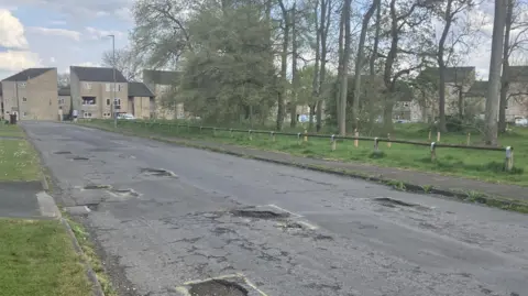 A road with several potholes on it. There are flats in the frame and trees with no leaves on them.