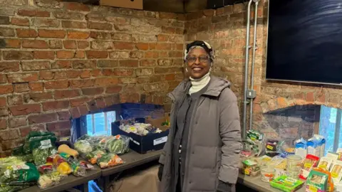 BBC/Tim Dale Martina O'Dwyer is wearing a headscarf and a grey long coat and stands in York's Red Tower surrounded by tables with food on them