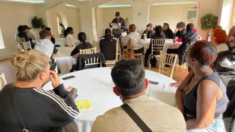 Un grupo de personas se sientan alrededor de mesas cubiertas con manteles blancos en una gran sala de Newstead Abbey. Las paredes son de color crema y la habitación está muy iluminada. 