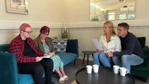 BBC Two pairs - two women and a man and a woman - are sat on two couches across each other in a room. They are looking at sheets of paper. There are white cups on the table in front of them. There is a mirror on the wall behind them.