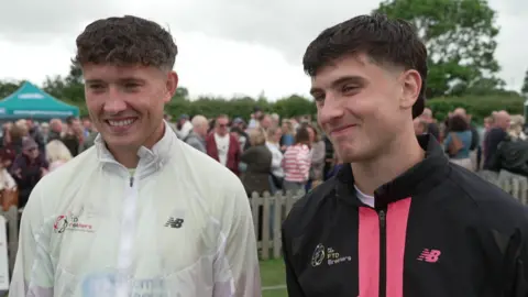 Two young men with short dark hair, one wearing a white running top and the other a black and pink running top