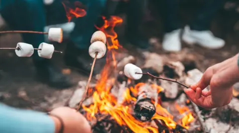 Getty Images A close up picture of people melting marshmallows on twigs on camp fire. There are logs burning on the fire and bright orange flames dancing around them. 