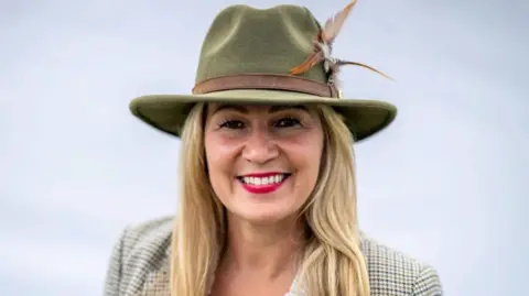 Getty Images Laura Anne Jones smiling looking at the camera. She is standing in front of a white background. Laura is wearing a beige tartan blazer and a wide-brimmed green hat. She has long blonde hair and is wearing bright pink lipstick. 