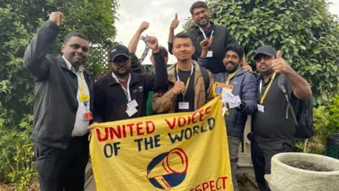 Seven members of the cleaning and porters team at St Helier and Epsom Hospital hold up a yellow banner with the name of their union, United Voices of the World, printed on it, as it each member of the group raises their hands in salute.