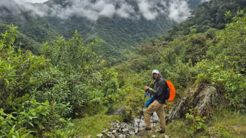 Jack Hazeldine Foundation Craig Plowman in walking gear and with poles on a steep jungle path surrounded by slopes covered in greenery and with low white clouds scudding by overhead. Craig is wearing a grey top, mustard-coloured cargo trousers and a white cap and he is carrying a bright orange backpack. He is smiling at the camera.