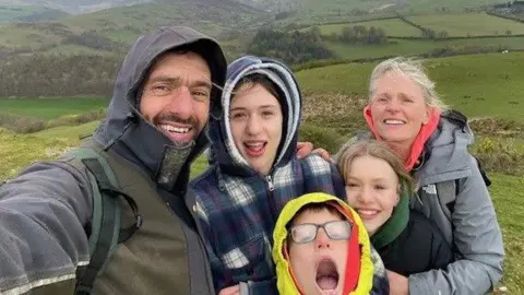 Rhodri Lloyd-Williams Mr Lloyd-Williams is smiling broadly as he takes a selfie with his family against rolling green hills. They wear coats with hoods up to shield from the elements.
