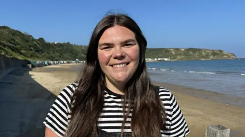 Mared Llywelyn wearing stripey top, standing in front of a beach looking at the camera