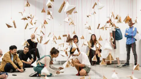 Sonica An art installation, which sees paper and wooden bird sculptures hanging down. Several people are examining them and looking at them. 