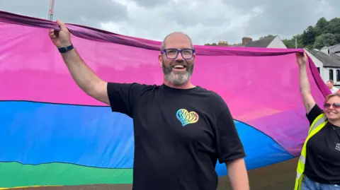A bald man wearing blue glasses smiles. He is wearing a black t-shirt and is holding the edge of a large flag, which is purple, blue, green, and yellow.