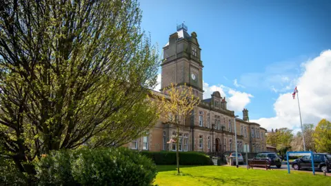 Historic looking cream-coloured building with a tall clock tower, viewed from greenery in front. A large leafy tree fills the left, while smaller flowering trees and shrubs frame the right. It is sunny and the sky is blue with a few clouds. Cars are parked near the entrance at the front of the building.