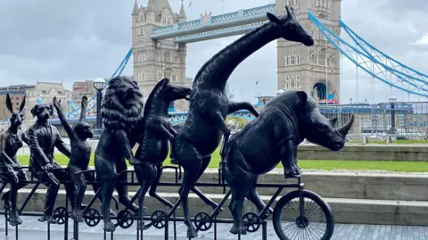 A metallic statue of a rhinoceros, giraffe, lion, monkey, dog and hare on a tandem bicycle. In the background is London's Tower Bridge on a cloudy day.