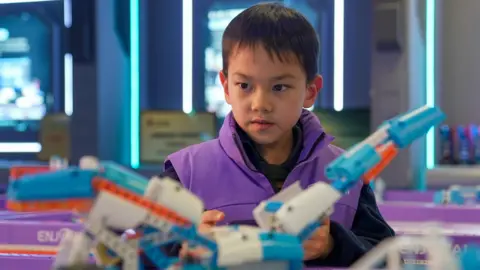 A child in a purple jacket operates a blue and orange AI-powered toy he built using code. 