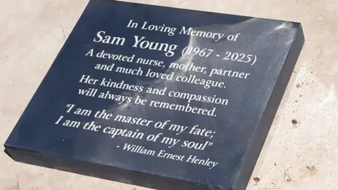 LDRS A grey plaque, inscribed with white lettering reading Sam Young, 1967-2025. A devoted nurse, mother, partner and much-loved colleague. Her kindness and compassion will also be remembered. "I am the master of my fate; I am the captain of my soul" - William Ernest Henley.