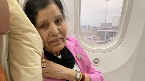 A woman with dark hair and a pink jacket in a white aeroplane seat with a window behind her