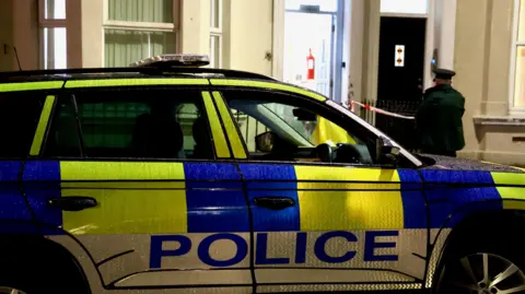 A police car outside a cream house. The door is open and an officer standing outside. The car is green white and blue.