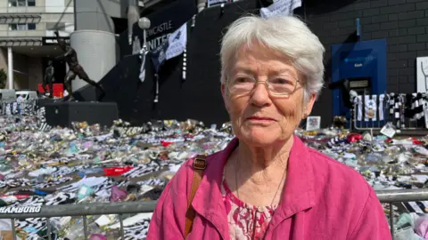 BBC/Kay Davison Anne Pontin standing outside of St James' Park which is still lined with Kevin Keegan tributes which spill out onto the pavement in front of the stadium. Pontin was short, white hair and is wearing silver rectangular glasses and a pink patterned top and shirt. She is looking to the left and has a straight face.