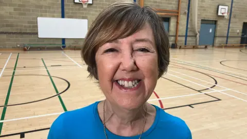 Christine is wearing a blue t-shirt, she has a small chain around her neck. She has short brown hair. She is standing in a sports hall. 