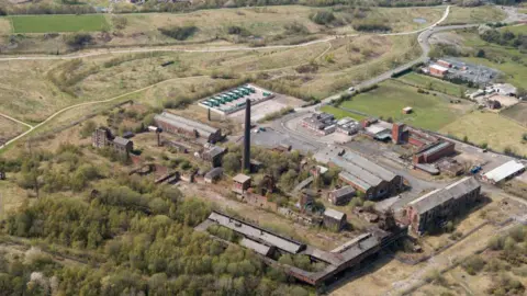 An aerial shot of the colliery site. It is a large area with concrete routes and grass, with steel buildings that are long. There are some large brown buildings and a tall chimney in the middle of the site.
