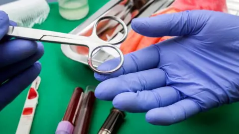 Getty Images Two blue-gloved hands pass a pair of metal surgical scissors over a green sterile operating drape. Nearby are several medical items including blood collection tubes with coloured caps, medical instruments arranged on a tray.