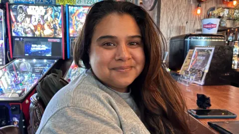 BBC News/Eloise Alanna Zeileen Reardon, wearing a grey sweatshirt, with her long dark hair loose, smiles at the camera. 