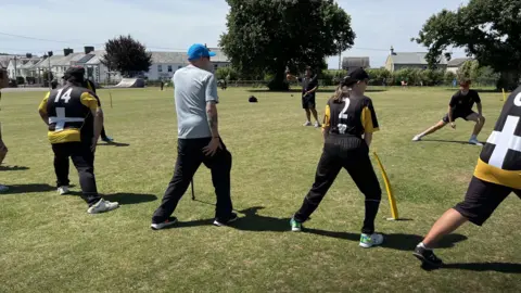 Five men and one woman are on a cricket pitch doing warm-up exercises and stretches. Five of them are wearing a sports strip which are black, gold and yellow with St Piran flags and a team number on them.