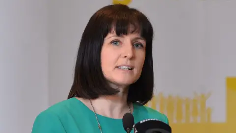 Pacemaker Alliance assembly member Paula Bradshaw, she had dark shoulder length hair and has a fringe.
She is wearing a green dress and is speaking into a microphone.