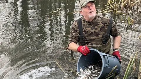 A man wearing camouflage gear holds a bucket of fish just above a river, ready to pour it in 