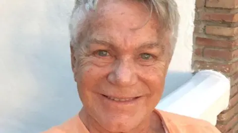 Family photograph Pierre Alexandre smiles for the camera. He is wearing an orange t-shirt, has blue eyes and cropped silver hair. 